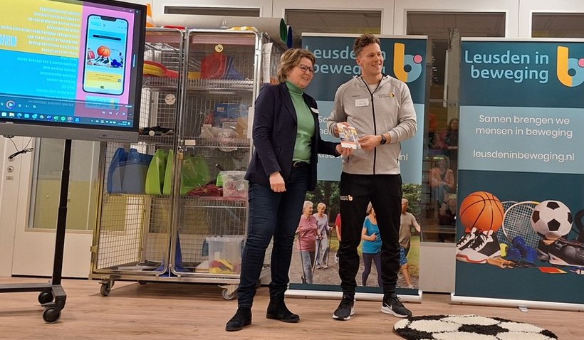 Marleen Treep, wethouder Sociaal Domein, neemt de nieuwe Max Vitaal-beweeggids in ontvangst van buurtsportcoach Tom tijdens het Sportcafé bij school De Atlas.